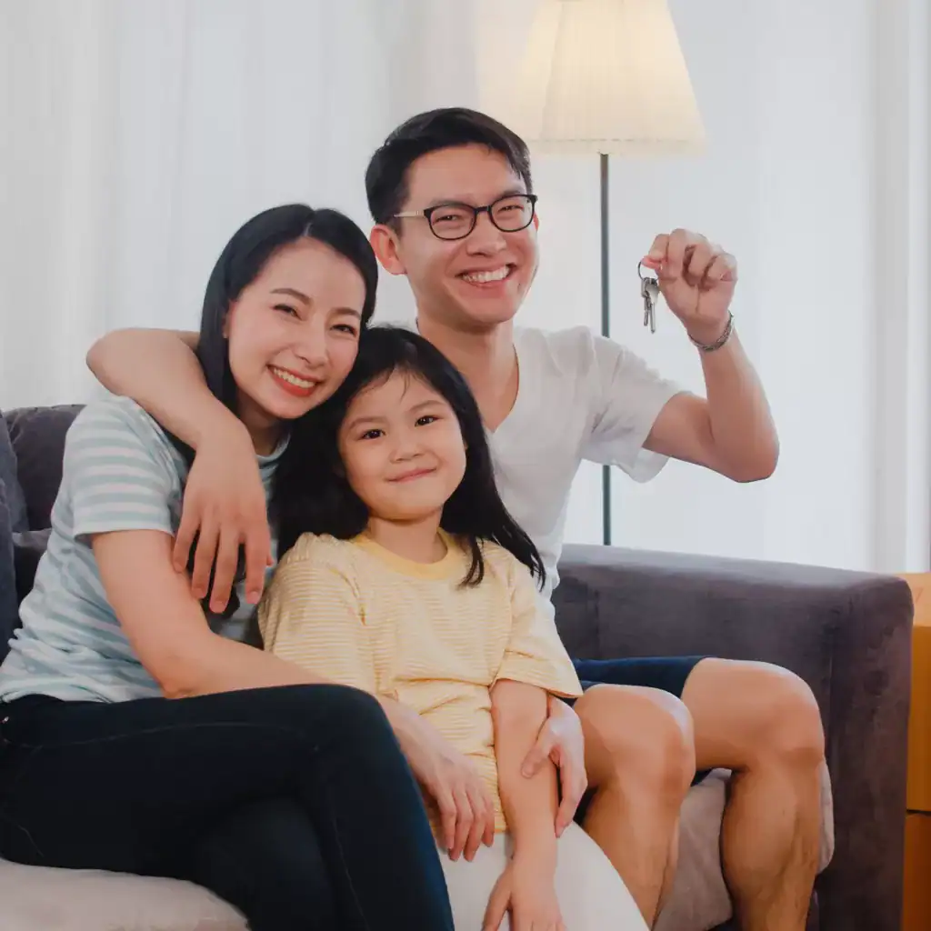 Portrait of Happy Asian young family bought new house. Japanese little preschool daughter with parents mother and father holds in hand keys sitting on sofa in living room smiling looking at camera.