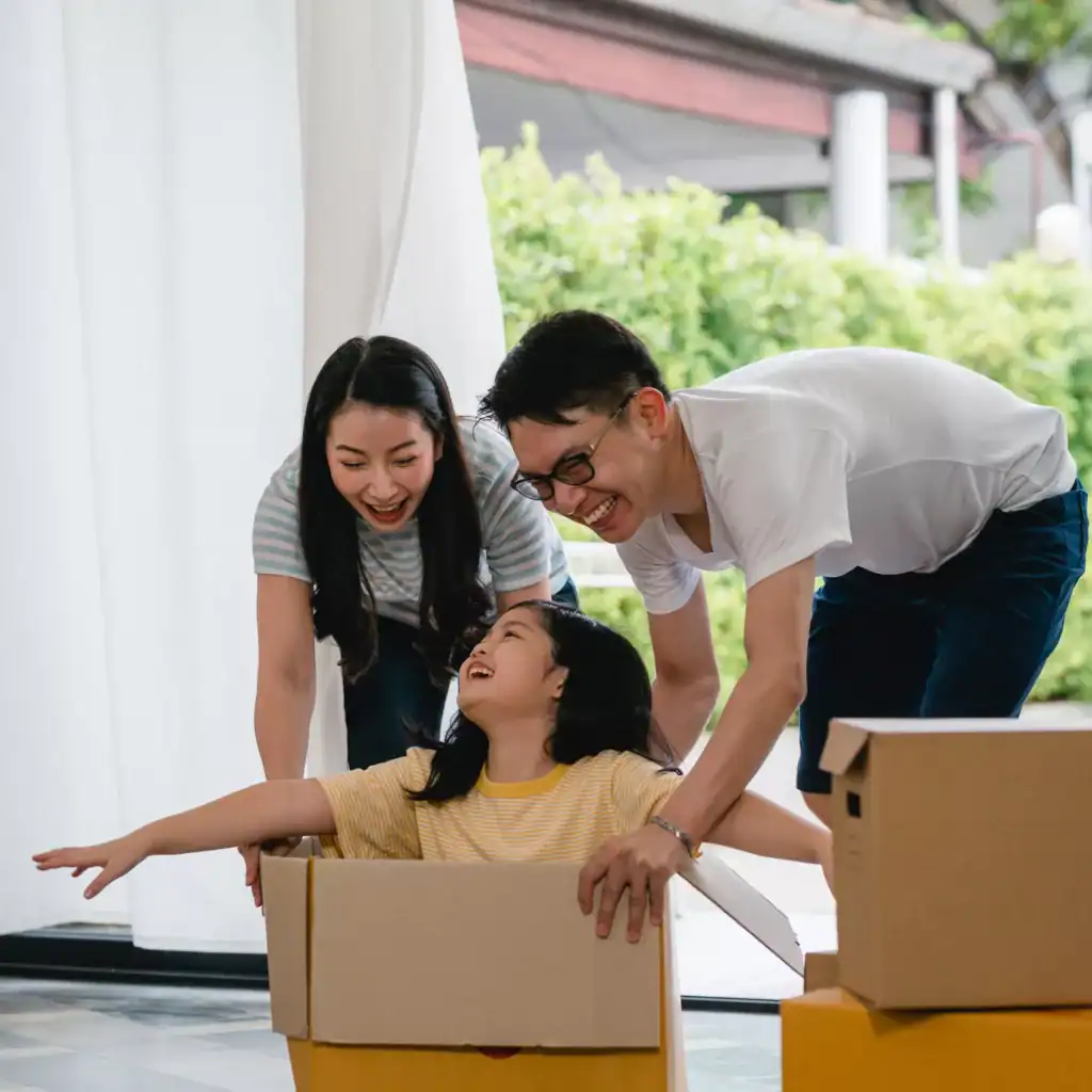 Happy Asian young family having fun laughing moving into new home. Japanese parents mother and father smiling helping excited little girl riding sitting in cardboard box. New property and relocation.